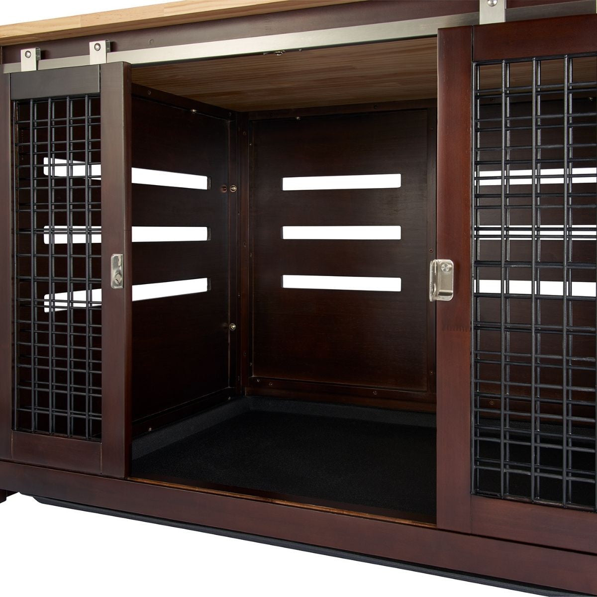 A close-up shot shows the open sliding doors of a dark brown wooden pet crate with black metal bars on the sides. The interior of the crate has a black mat on the floor and horizontal ventilation slots on the side walls.