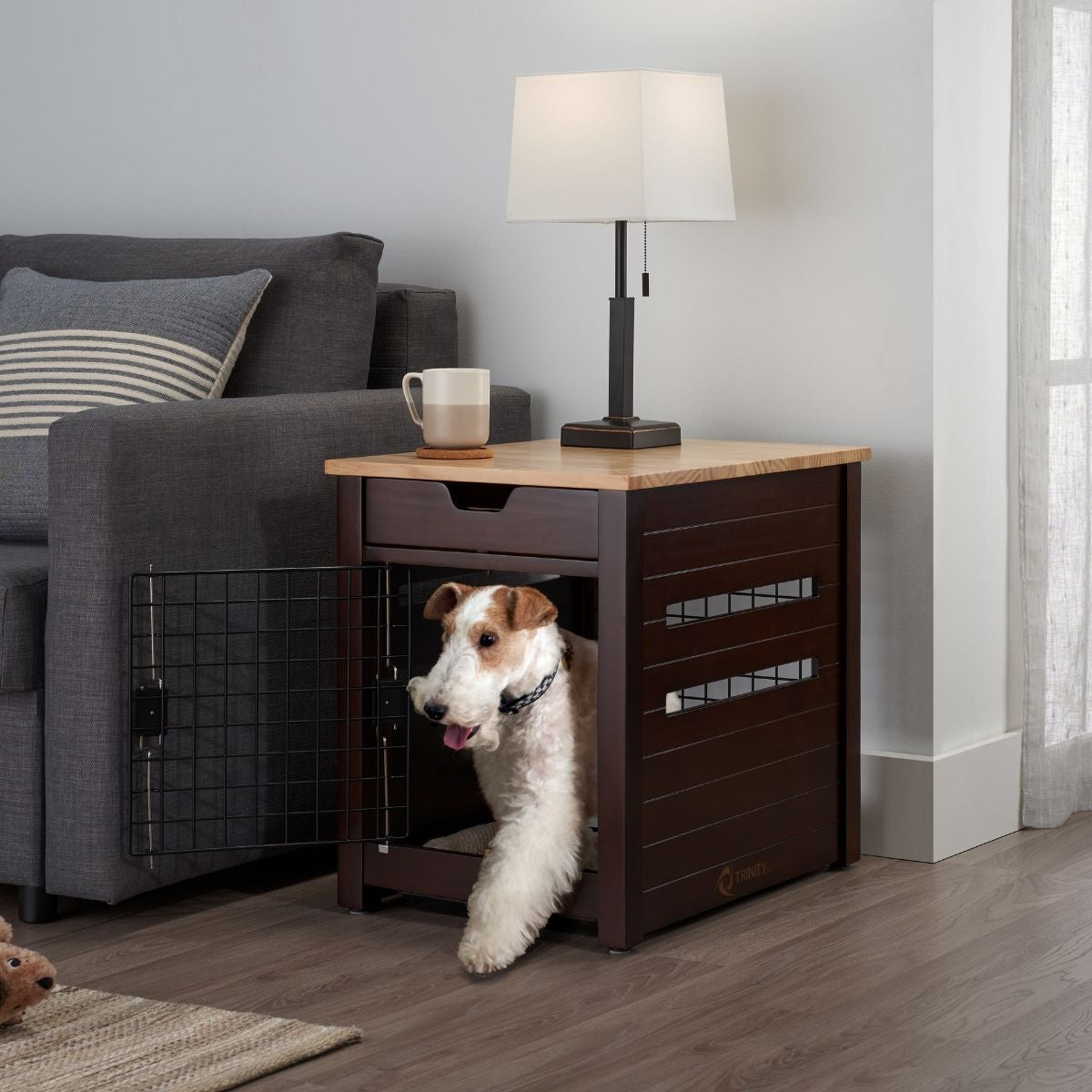 A medium-sized, white and brown wire fox terrier dog with its tongue slightly out stands in an open brown wooden dog crate that doubles as an end table next to a gray couch. A white mug sits on a wooden coaster on the end table under a white lampshade.