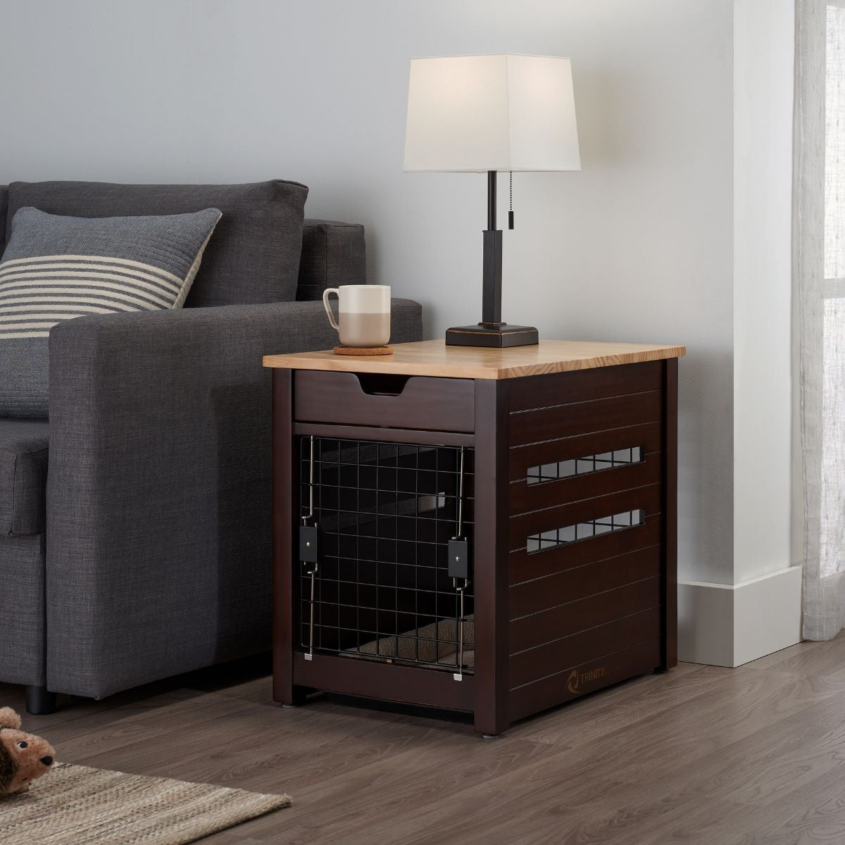A dark brown wooden pet crate doubles as an end table next to a gray couch. A white lamp with a square shade and a beige mug sit on the wooden tabletop.