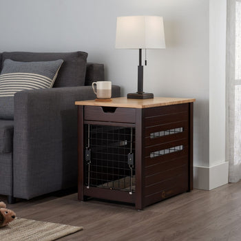 A dark brown wooden pet crate doubles as an end table next to a gray couch. A white lamp with a square shade and a beige mug sit on the wooden tabletop.
