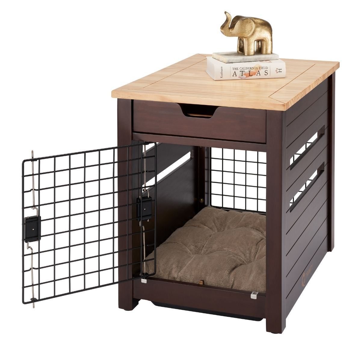 A brown wooden pet crate doubles as an end table, featuring a wire mesh door that is currently open. The top of the crate, made of lighter-colored wood, holds a book titled "ATLAS" and a small gold-colored elephant figurine.