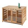 A light wood pet crate doubles as an accent table, featuring sliding barn-style doors with black metal mesh and hardware. A green water bottle and a black bone-shaped toy rest on the top surface.