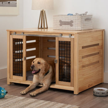 A golden retriever dog rests inside a light wood pet crate that doubles as an accent table. The crate has sliding metal-framed doors with black wire mesh and is topped with a lamp and a fabric storage bin with a bone design.