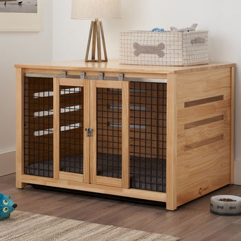 A light wood pet crate doubles as an accent table, featuring sliding metal mesh doors with a latch. On top, a lamp and a fabric storage bin with a bone design sit, while a pet bowl rests on the floor nearby.