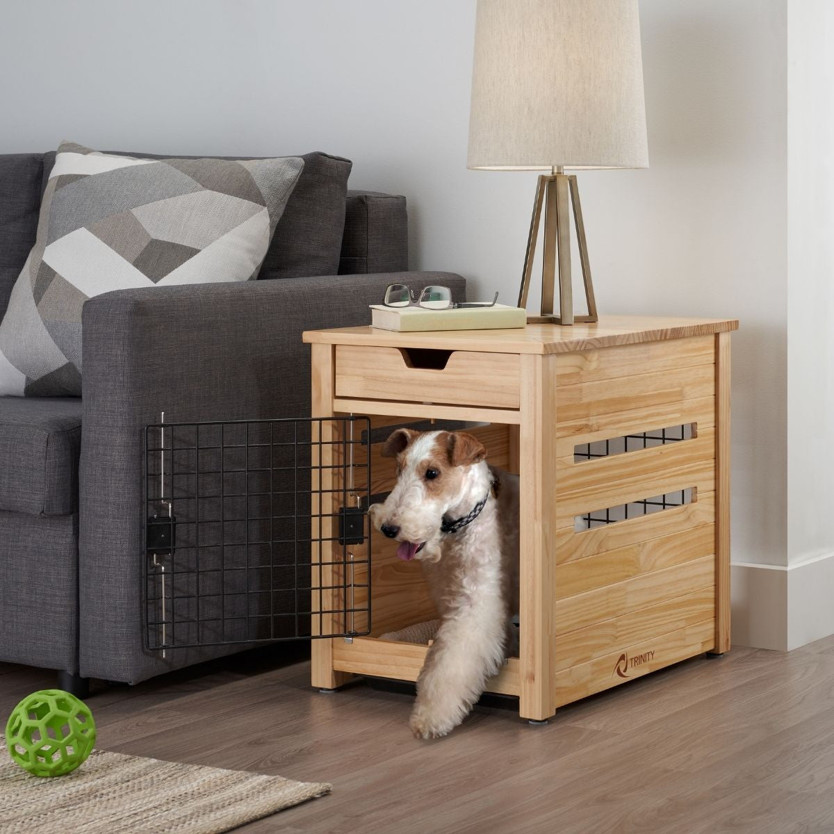 A medium-sized, white and brown terrier dog with its tongue sticking out stands inside a light-colored wooden dog crate that doubles as an end table. The crate has a black wire door that is partially open, and it sits next to a gray couch with a patterned pillow.