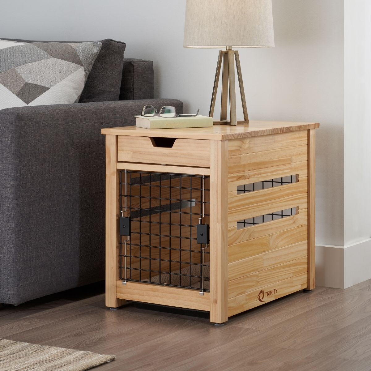 A light wood end table functions as a dog crate, featuring a black wire mesh door, a drawer, and horizontal slats on the sides for ventilation. The table sits on a dark wooden floor next to a gray couch with patterned pillows and under a lamp with a light-colored shade.