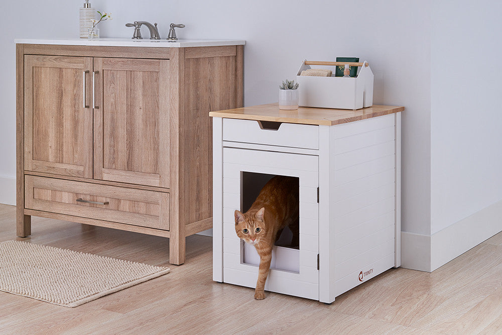 A ginger cat is stepping out of a white wooden enclosure designed to look like a small cabinet, placed beside a larger wooden bathroom vanity. The enclosure has a smooth wooden top with a small caddy and plant placed on it, blending seamlessly into the clean, modern decor.