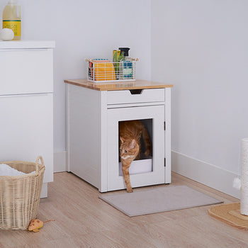 A white cat exits a white wooden cat litter box enclosure with a drawer, positioned in a corner of a room with light-colored wood flooring. The enclosure has a bamboo top, and a woven basket and cleaning supplies are visible on top.