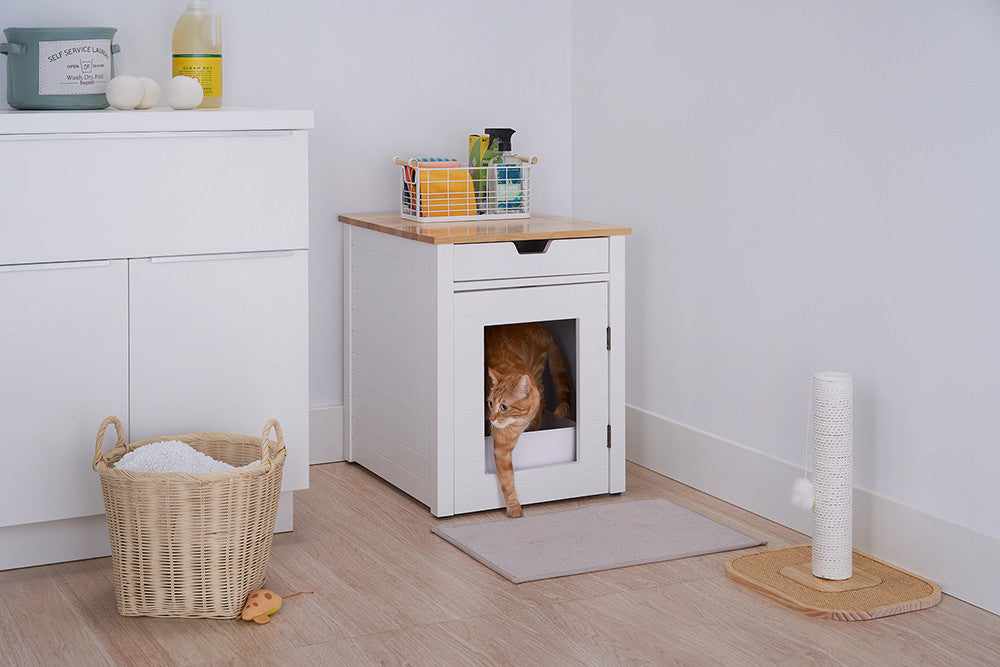 A white cat exits a white wooden cat litter box enclosure with a drawer, positioned in a corner of a room with light-colored wood flooring. The enclosure has a bamboo top, and a woven basket and cleaning supplies are visible on top.