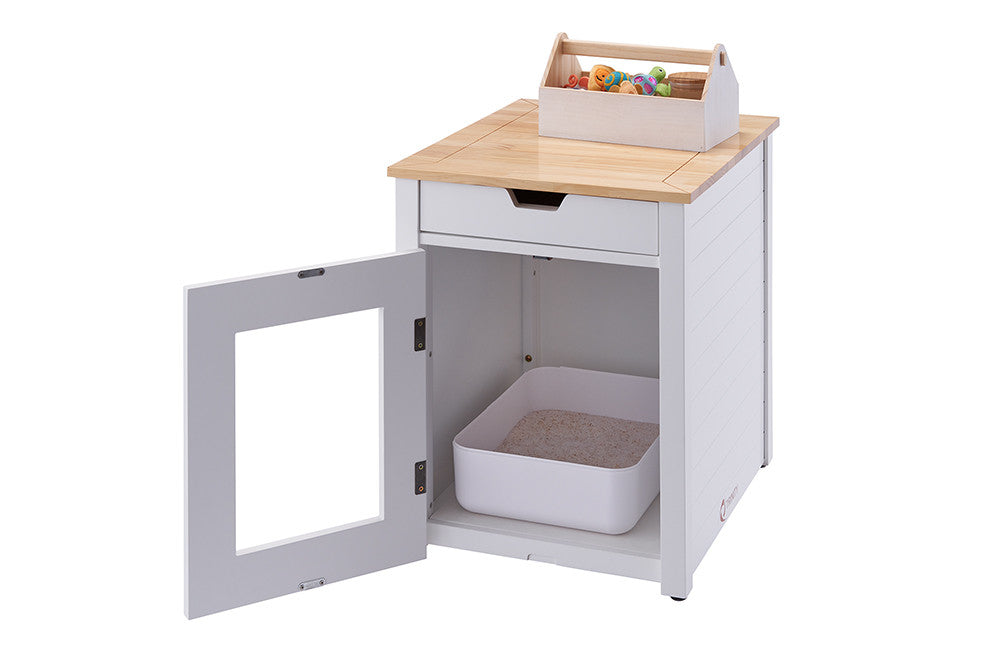 A white end table with a light wood top features an open door revealing a cat litter box inside. A small drawer is visible above the door, and a toy box sits on top of the table.