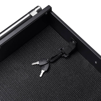 A top-down, close-up shot shows the interior of a black safe. Two silver keys attached to a black lanyard with a velcro strap rest on the safe's textured black bottom.