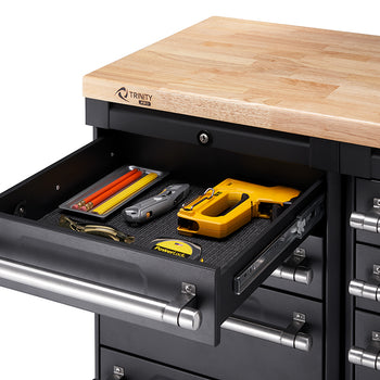 A close-up shot shows a black Trinity PRO toolbox with a wooden countertop. The top-most drawer is open, revealing a black textured liner holding several items including yellow and red pencils, a silver utility knife, and a yellow staple gun.