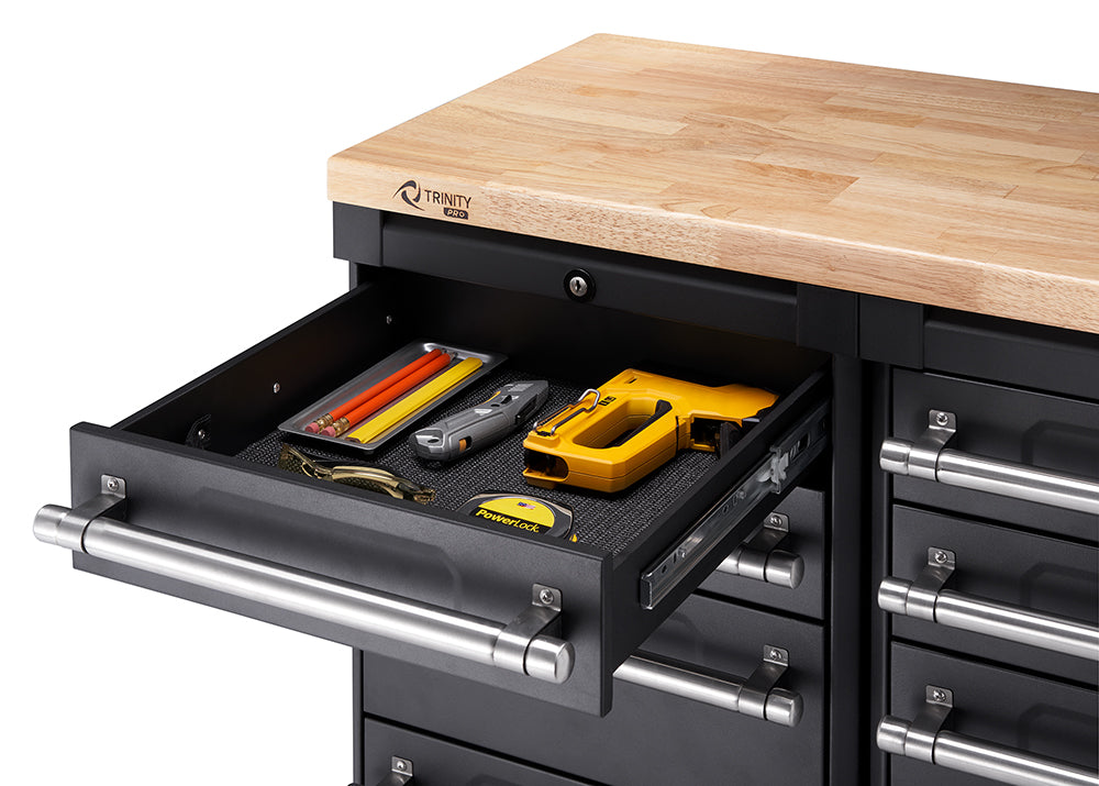 A close-up shot shows a black Trinity PRO toolbox with a wooden countertop. The top-most drawer is open, revealing a black textured liner holding several items including yellow and red pencils, a silver utility knife, and a yellow staple gun.