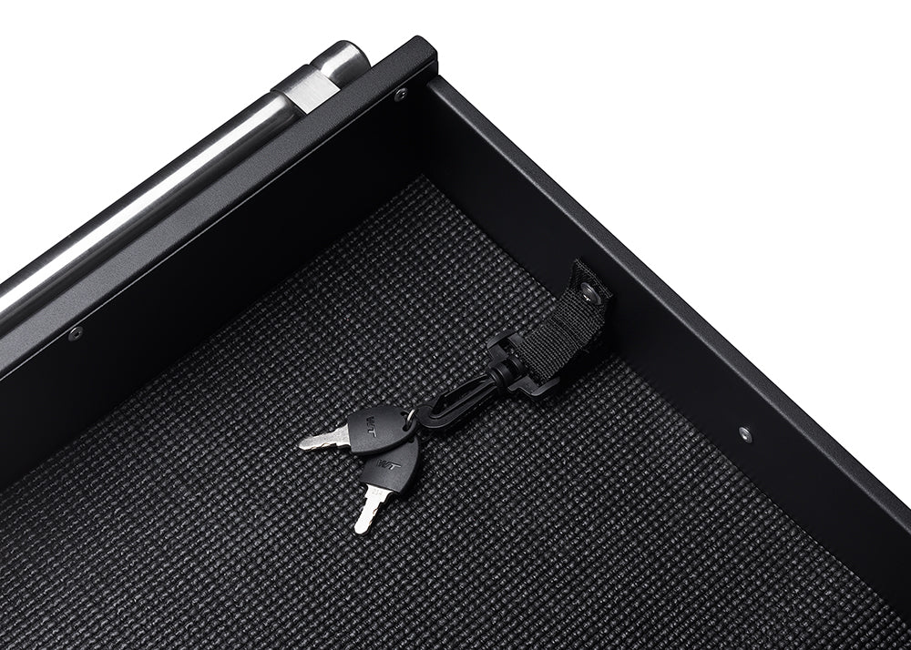 A pair of silver keys on a black lanyard are clipped to a black fabric loop inside a black drawer. The drawer has a textured, possibly rubber, bottom liner.