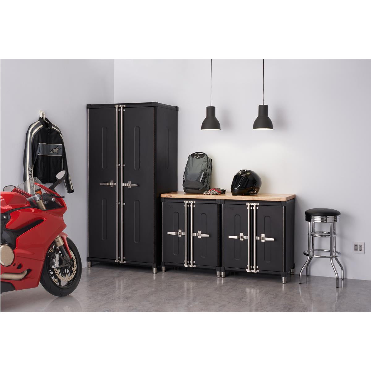 A section of a garage is shown with a four-piece black cabinet set against a light gray wall. A red motorcycle is parked to the left, and various garage-related items like a jacket, backpack, helmet, gloves, and a stool are arranged around the cabinets.