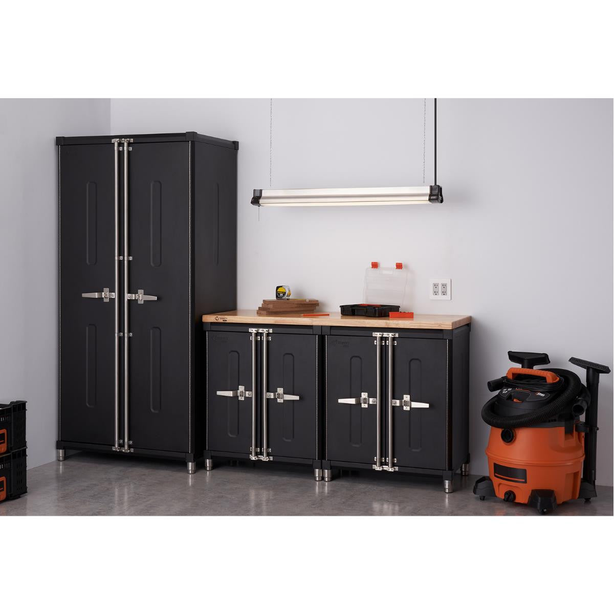 A well-organized garage corner features a tall, two-door black Trinity Pro Series cabinet on the left, and two shorter black cabinets with a shared light wood countertop to the right. An orange wet/dry vacuum cleaner stands to the right of the shorter cabinets.