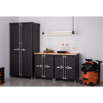 A well-organized garage corner features a tall, two-door black Trinity Pro Series cabinet on the left, and two shorter black cabinets with a shared light wood countertop to the right. An orange wet/dry vacuum cleaner stands to the right of the shorter cabinets.