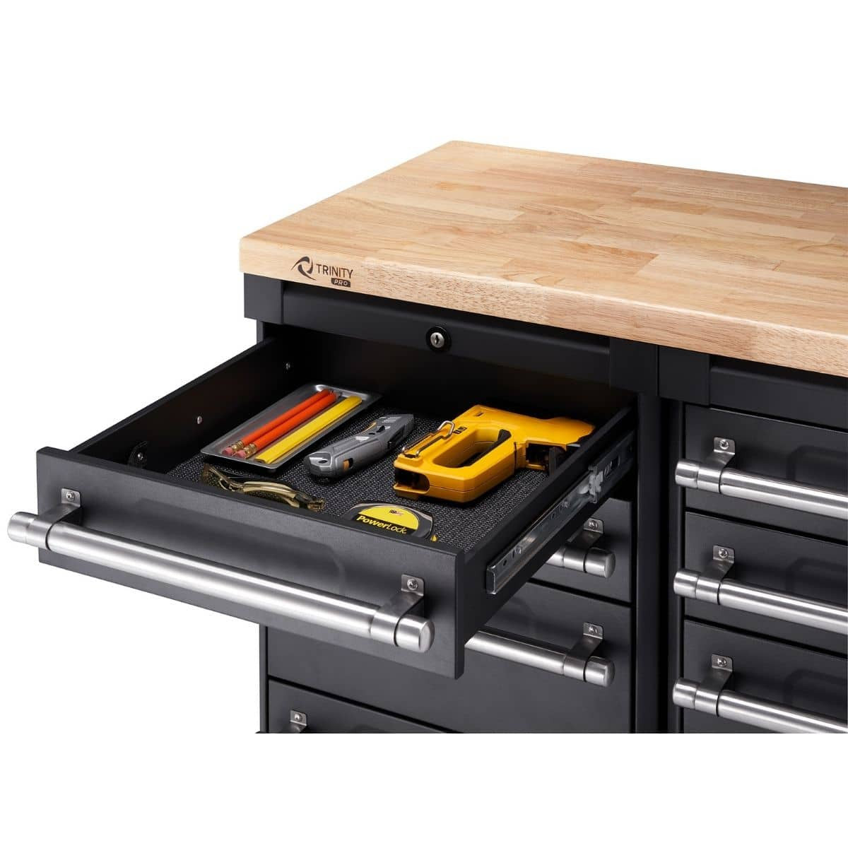 This close-up shot shows a partially opened drawer of a black Trinity Pro garage cabinet, revealing several tools neatly arranged inside on a black liner. The drawer has a stainless steel handle and contains items like pliers, a yellow staple gun, and red and yellow handled tools.