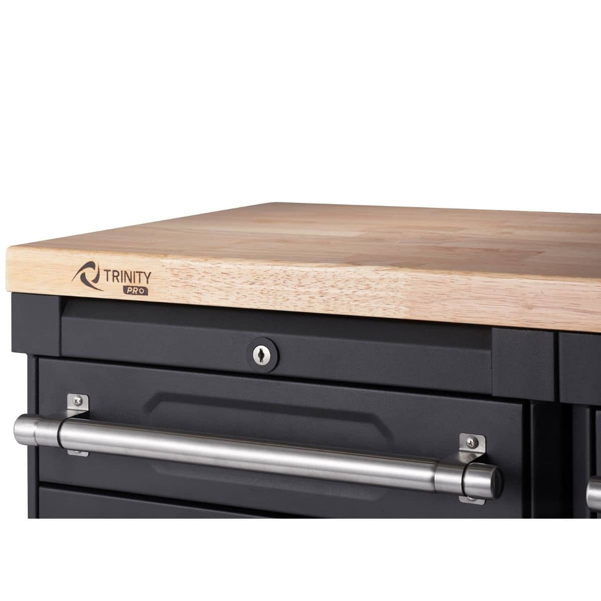 A close-up shows the top corner of a black Trinity Pro garage cabinet drawer set with a light-colored wooden countertop. A silver handle is attached to the front of the top drawer, and a keyhole lock is visible above it.