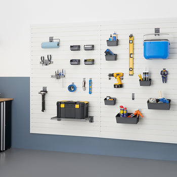 A garage wall features a white slatwall organization system holding various tools and supplies, including a cooler, toolbox, power drill, and hand tools. To the left, a black cabinet with a wooden top provides additional storage.