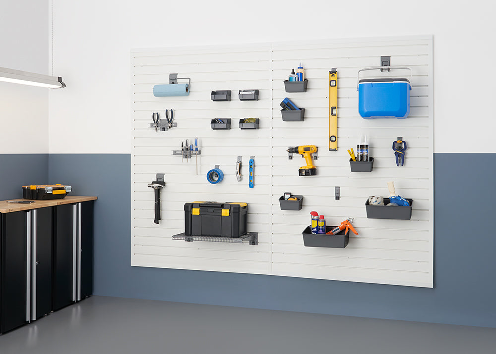 A garage wall features a white slatwall organization system holding various tools and supplies, including a cooler, toolbox, power drill, and hand tools. To the left, a black cabinet with a wooden top provides additional storage.