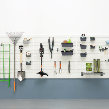 A well-organized garage wall features various gardening tools neatly hung on a slatwall system. The tools include rakes, shovels, trimmers, trowels, and gardening boots, along with bins for smaller items and a paper towel holder.