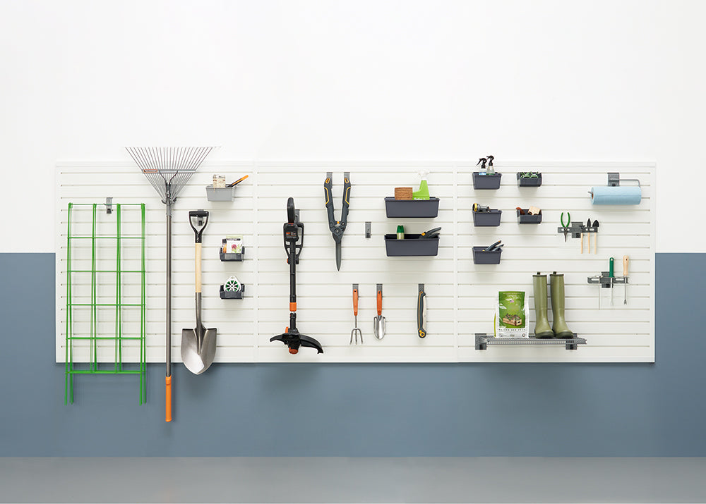 A well-organized garage wall features various gardening tools neatly hung on a slatwall system. The tools include rakes, shovels, trimmers, trowels, and gardening boots, along with bins for smaller items and a paper towel holder.