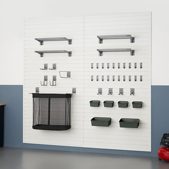 A clean and organized garage wall features a white slatwall system with various shelving, hooks, and bins holding tools and other items. To the left is a black cabinet with a wooden countertop, and on the right, a red air compressor sits on the floor.