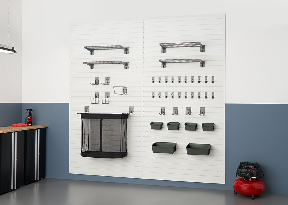 A clean and organized garage wall features a white slatwall system with various shelving, hooks, and bins holding tools and other items. To the left is a black cabinet with a wooden countertop, and on the right, a red air compressor sits on the floor.