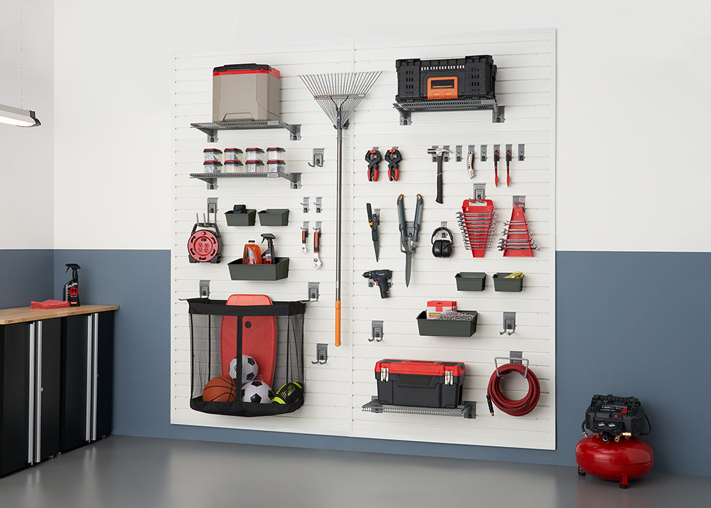 A neatly organized garage wall features white slatwall panels holding various tools, sports equipment, and storage containers. On the left, a black cabinet with a wooden top stands against a two-toned wall.