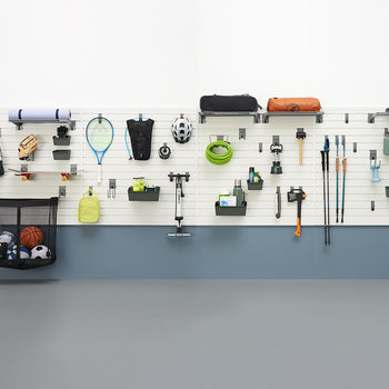 A garage organization system features a white slatwall holding various sports and outdoor gear. Items include baseball bats, a skateboard, tennis rackets, camping equipment, hiking poles, and a mesh bag filled with balls.