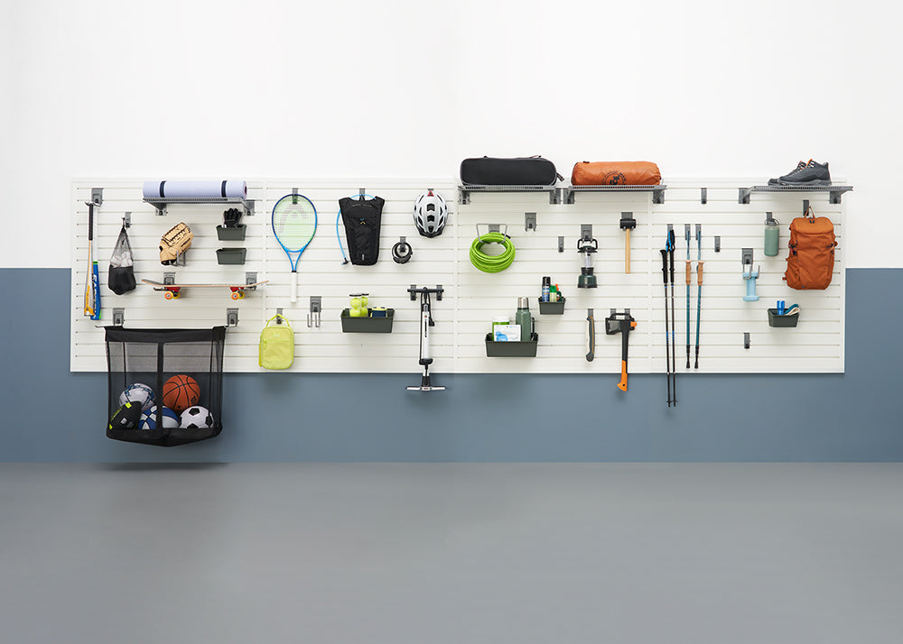 A garage organization system features a white slatwall holding various sports and outdoor gear. Items include baseball bats, a skateboard, tennis rackets, camping equipment, hiking poles, and a mesh bag filled with balls.