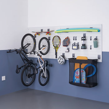 A well-organized garage wall storage system holds various sports and outdoor equipment, including two bicycles, a skateboard, a tennis racket, a baseball glove, a helmet, and a mesh bag filled with beach toys. The white slatwall panels with adjustable hooks and holders keep everything neatly arranged above a blue and gray painted floor.