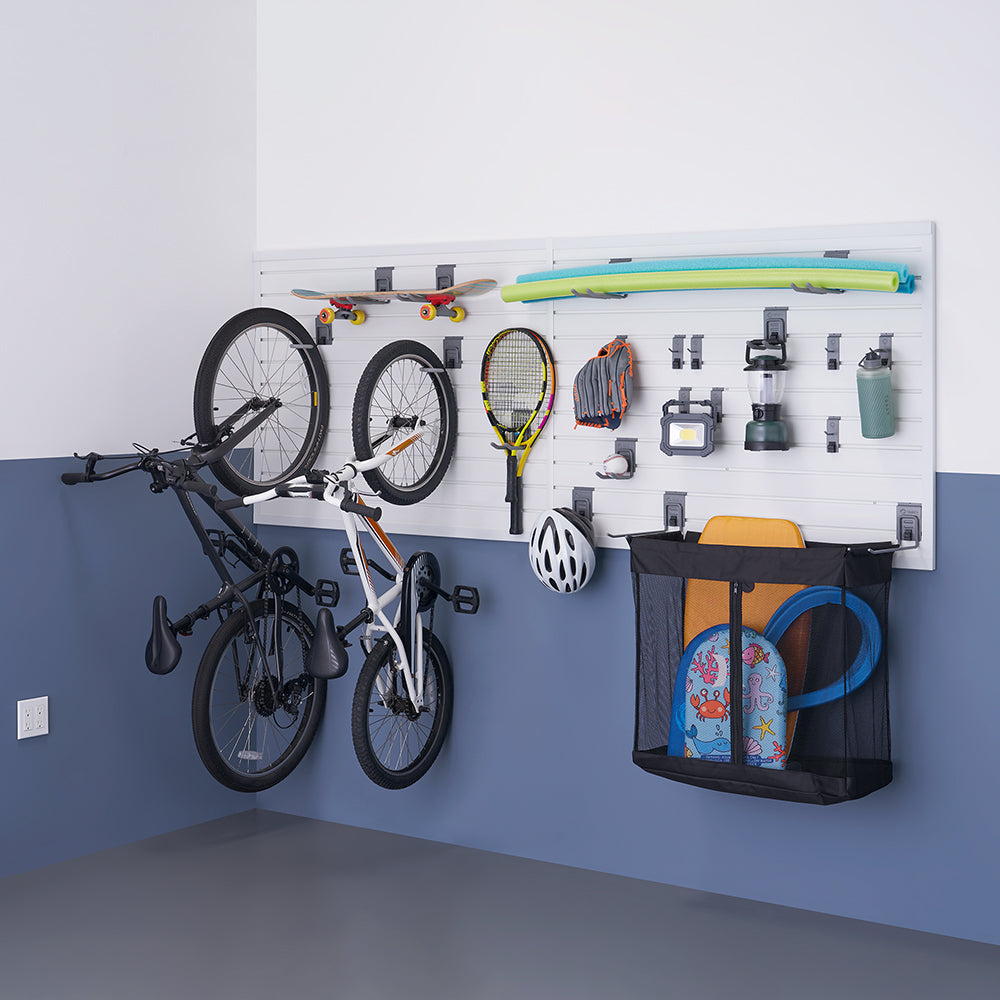 A well-organized garage wall storage system holds various sports and outdoor equipment, including two bicycles, a skateboard, a tennis racket, a baseball glove, a helmet, and a mesh bag filled with beach toys. The white slatwall panels with adjustable hooks and holders keep everything neatly arranged above a blue and gray painted floor.