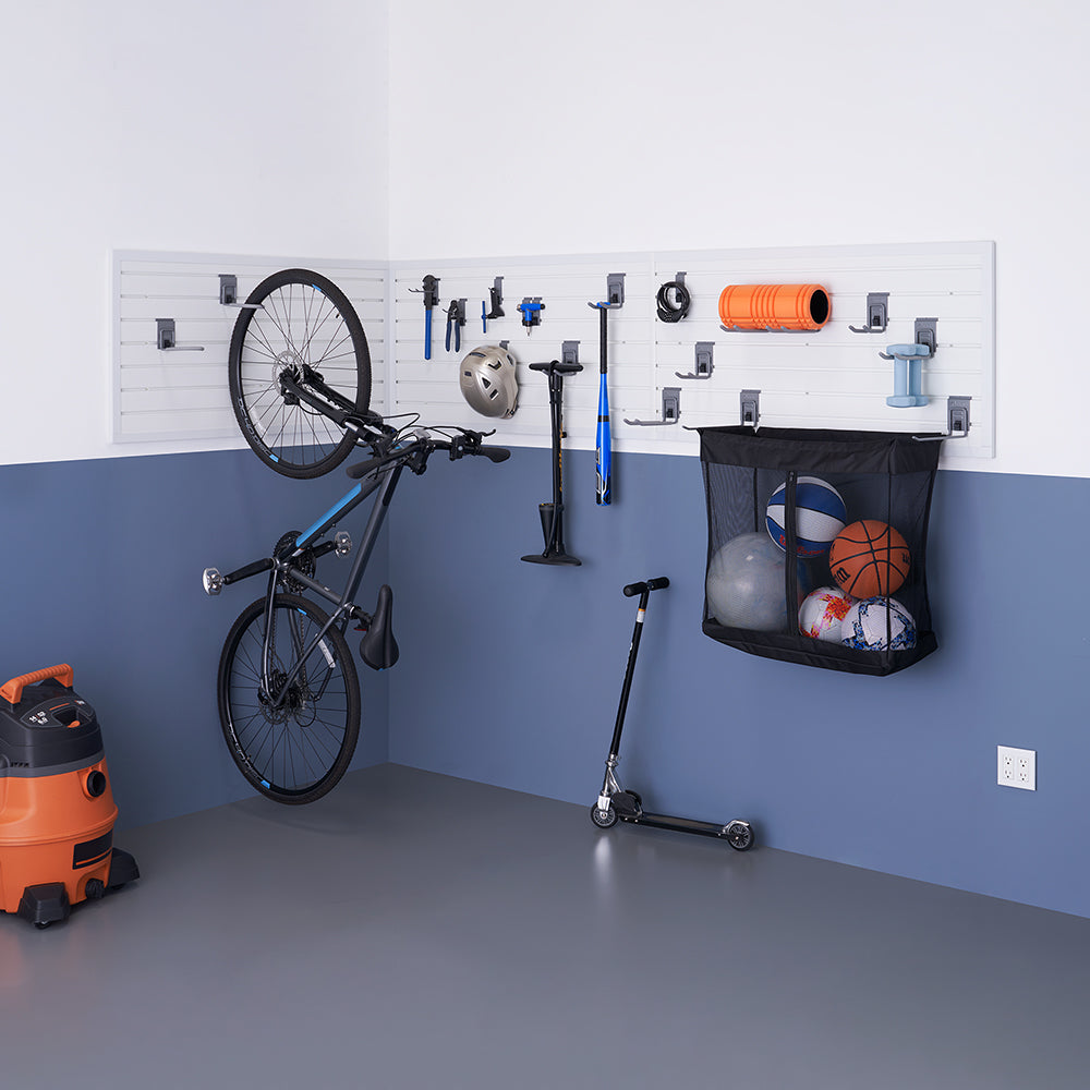 A well-organized garage storage system featuring a mounted slat wall with various hooks and holders for a bicycle, scooter, sports balls, tools, and accessories. The lower half of the wall is painted blue, and a vacuum cleaner is placed on the gray floor.