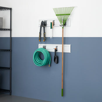 A storage wall system in a garage or shed displays various garden tools and a coiled green hose. A large black wire shelving unit stands to the left, holding various items including a terracotta pot.