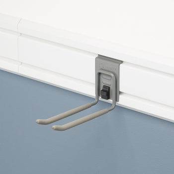 A close-up view of a gray, double-pronged hook mounted on a white slatwall panel. The hook has a spring-loaded lock and is designed for garage organization.