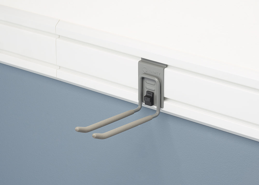 A close-up view of a gray, double-pronged hook mounted on a white slatwall panel. The hook has a spring-loaded lock and is designed for garage organization.
