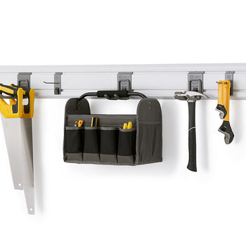 A hand saw, tool bag, hammer, and wrench are hanging from hooks on a white slatwall panel. The panel is mounted on a white wall in what appears to be a garage or workshop, providing an organized storage solution for various tools.