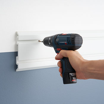 A close-up shot shows a person's hand using a blue and black Bosch cordless drill to screw a screw into a white plastic wall-mounted storage rail. The wall is painted white on top and a slate blue on the bottom, with the storage rail installed at the color transition line.