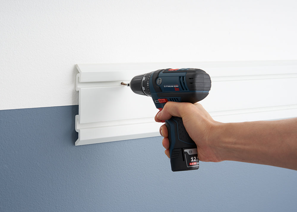 A close-up shot shows a person's hand using a blue and black Bosch cordless drill to screw a screw into a white plastic wall-mounted storage rail. The wall is painted white on top and a slate blue on the bottom, with the storage rail installed at the color transition line.