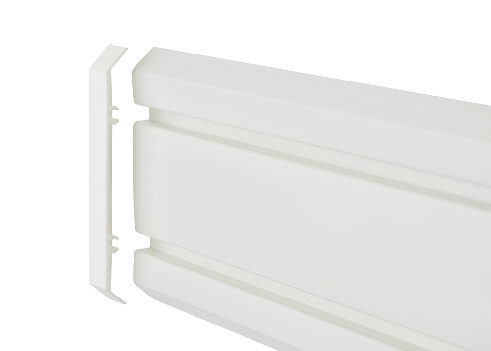 A detailed, eye-level close-up shot shows a white, horizontally-oriented plastic object, likely a radiator cover or a plinth heater. A separate, thin, flat end-cap piece is shown detached and aligned with the left end of the main object, revealing how it would fit together.
