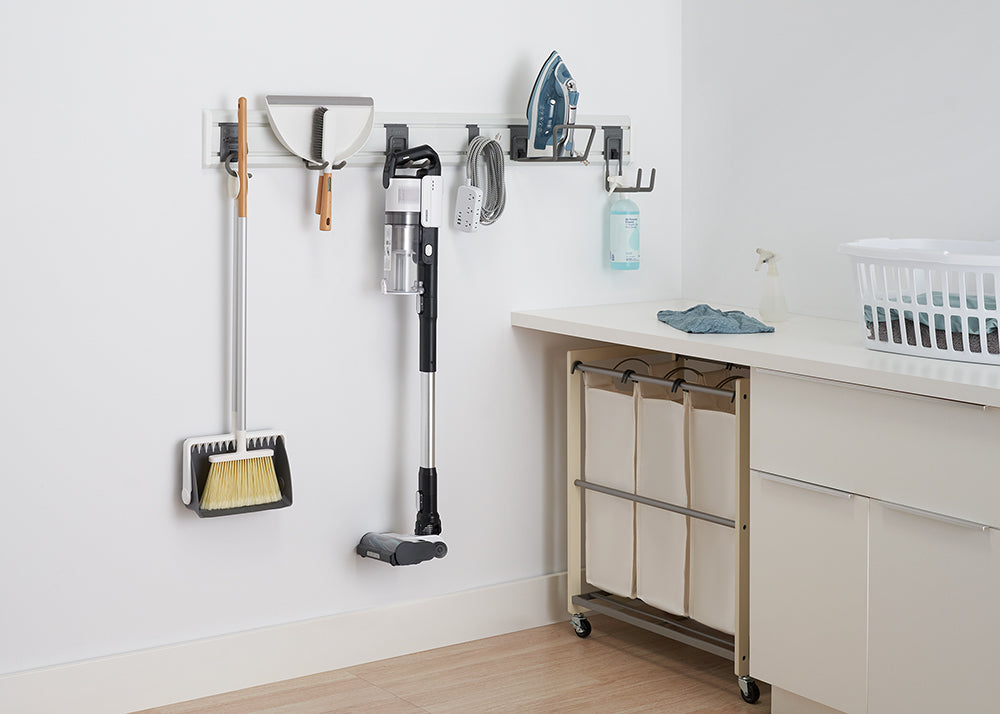 A wall-mounted organization system in a laundry room holds a broom, dustpan, and cordless vacuum. An iron and other cleaning supplies hang nearby, while a rolling laundry sorter is tucked under a white countertop.