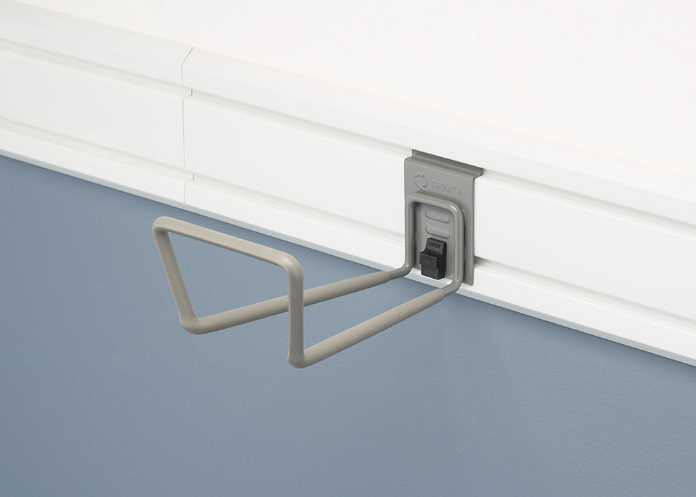 A close-up shot of a gray TRINITY brand slat wall hook mounted on a white slat wall. The hook has two prongs shaped like a flattened "U" and a black locking mechanism to secure it to the wall.