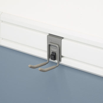 A close-up shot of a gray, J-shaped hook attached to a white slatwall panel. The hook is from the brand TRINITY, and is designed to hang tools or other items on the wall.