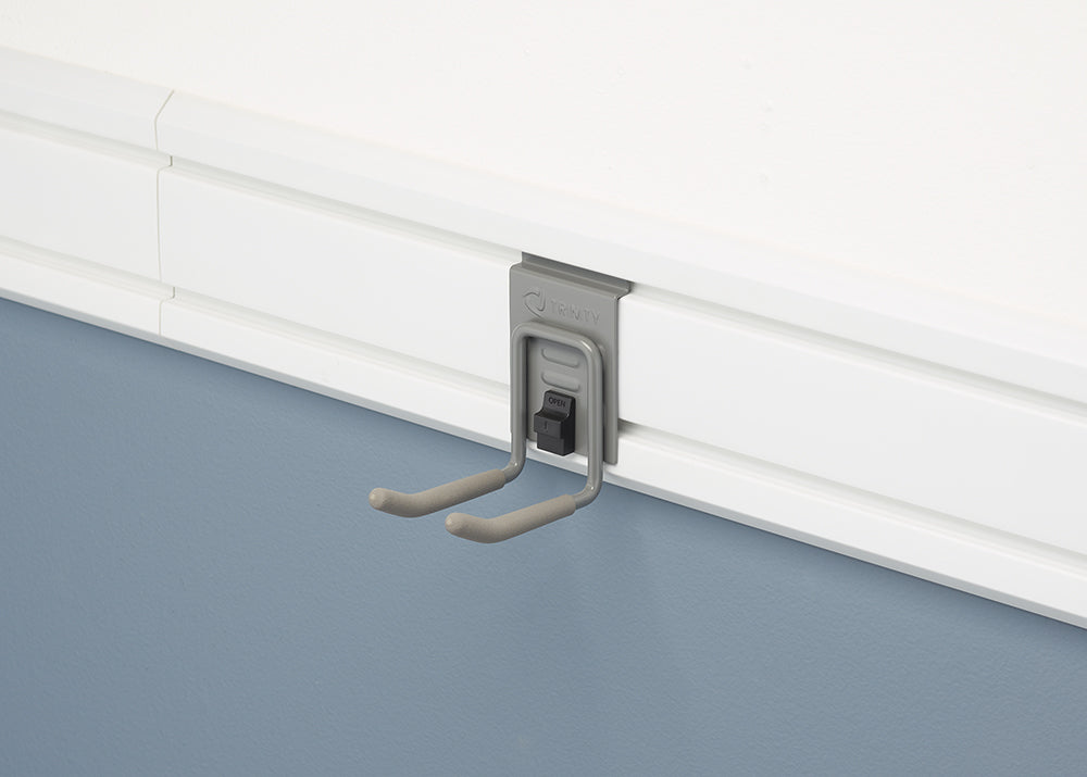 A close-up shot of a gray, J-shaped hook attached to a white slatwall panel. The hook is from the brand TRINITY, and is designed to hang tools or other items on the wall.