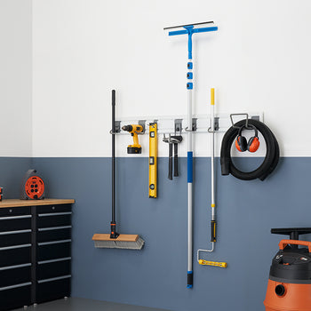 A clean and organized garage wall with a variety of tools hanging on a storage rack. The tools include a broom, a drill, a level, a window squeegee, a paint roller, and a shop vacuum with headphones hanging on it.