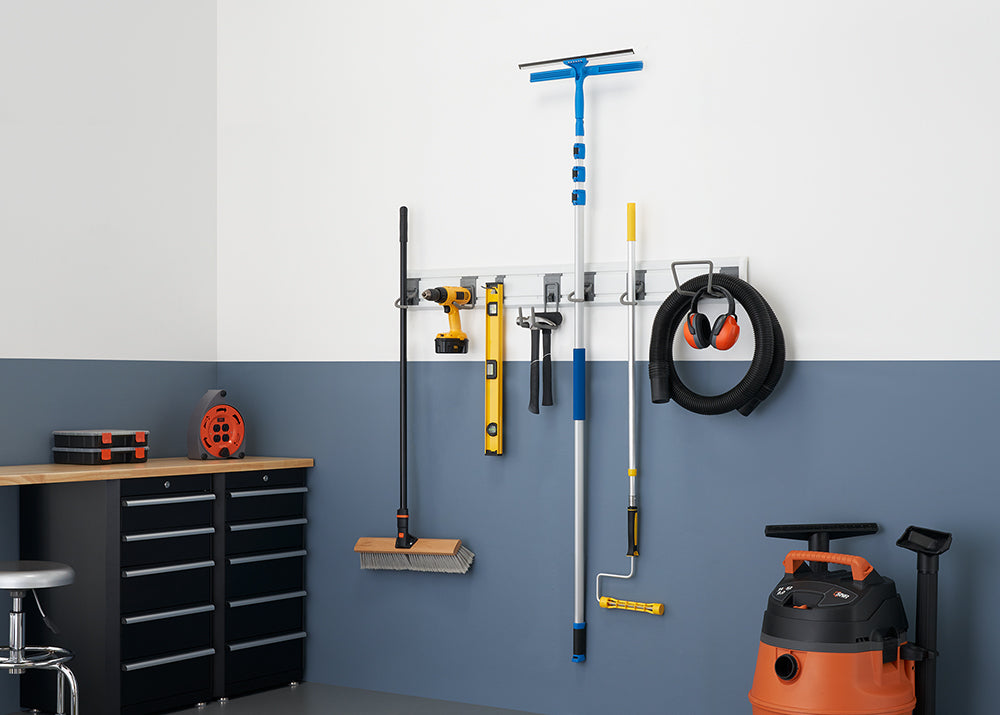 A clean and organized garage wall with a variety of tools hanging on a storage rack. The tools include a broom, a drill, a level, a window squeegee, a paint roller, and a shop vacuum with headphones hanging on it.