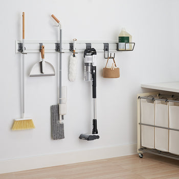 A white wall with a mounted storage system holding various cleaning supplies. Items include a broom, dustpan, mop, vacuum cleaner, and a hanging basket with a cleaning solution bottle and a small basket.