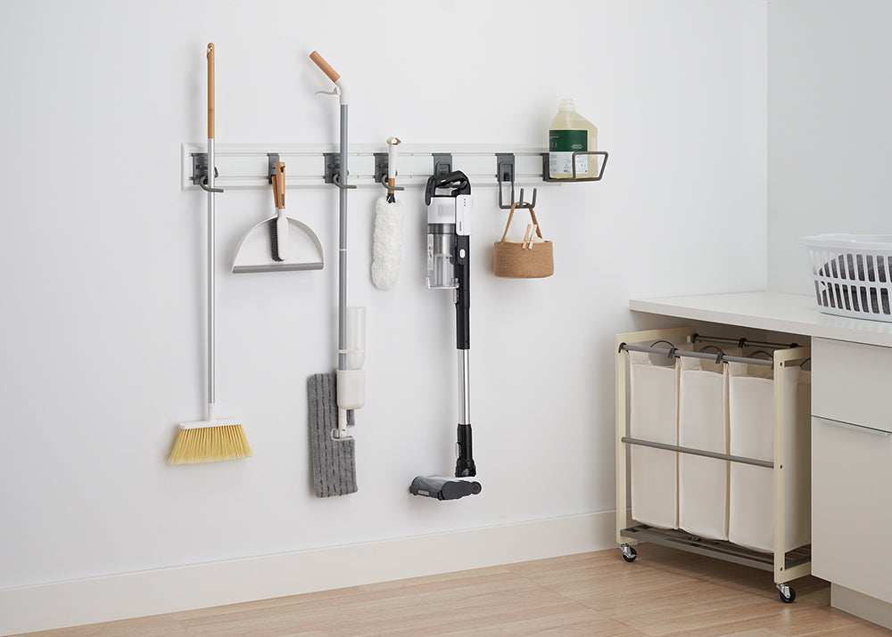 A white wall with a mounted storage system holding various cleaning supplies. Items include a broom, dustpan, mop, vacuum cleaner, and a hanging basket with a cleaning solution bottle and a small basket.